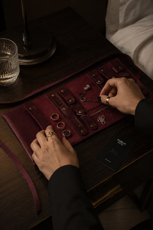 Burgundy leather jewelry organizer unfolded flat on a nightstand, showing earring holders and a ring bar for travel